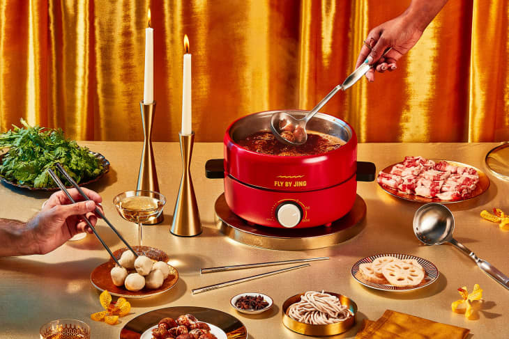 Red hot pot with broth, surrounded by plates of meat, vegetables, noodles, and lit candles on a gold table.
